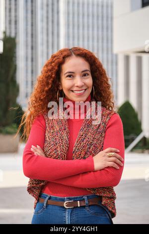 Eine stilvolle Lateinfrau mit lockigem Haar steht selbstbewusst in einer urbanen Umgebung, trägt einen roten Pullover und eine farbenfrohe Weste Sie lächelt warm und verkörpert ein V Stockfoto