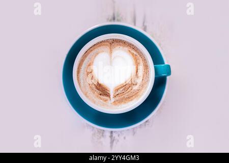 Ein wunderschön gefertigter Cappuccino mit herzförmigem Schaumstoffdesign in einer leuchtend blauen Tasse. Die komplizierte Latte Art verleiht einem Hauch von Liebe und Kreativität Stockfoto