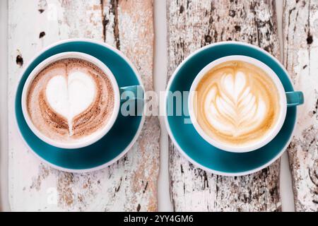 Zwei blaue Tassen mit Cappuccino und heißer Schokolade mit Herz- und Blatt-Latte-Art sitzen auf einem verwitterten Holztisch und bieten einen gemütlichen, rustikalen Kaffee-Sho Stockfoto