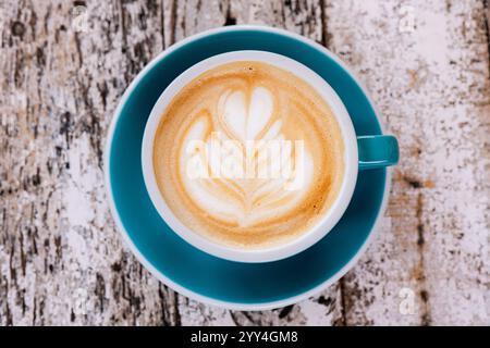 Blick von oben auf einen Cappuccino in einer blauen Tasse mit komplizierter Latte Art auf einem rustikalen Holztisch. Die ruhige Komposition unterstreicht den Kaffeekultu Stockfoto