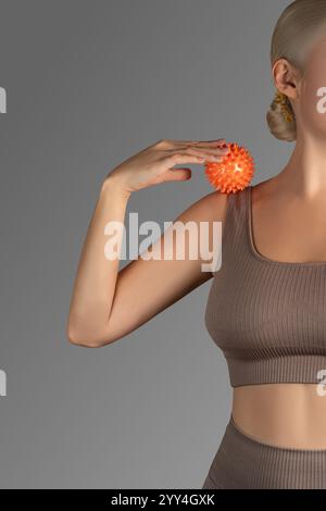 Eine nicht erkennbare Frau, die einen leuchtenden, orangefarbenen, stacheligen Massageball auf der Schulter verwendet, demonstriert eine Fitness- und Selbstpflegeroutine. Sie ist angezogen Stockfoto