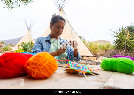 Eine Frau konzentriert sich darauf, im Freien ein buntes Garn zu kreieren. Sie sitzt an einem Holztisch mit leuchtenden Garnbällen und Tipis im Hintergrund, enjo Stockfoto