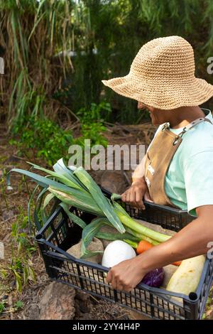 Ein Landwirt trägt einen Strohhut und eine Schürze mit frisch geerntetem Gemüse, einschließlich Lauch, Zucchini, Zwiebeln und Karotten, in A Stockfoto