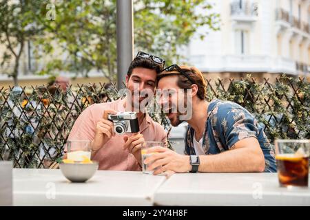 Ein fröhliches schwules Paar sitzt draußen an einem Café-Tisch und lacht, während es Fotos auf einer alten Kamera ansieht und einen sonnigen Tag genießt. Stockfoto