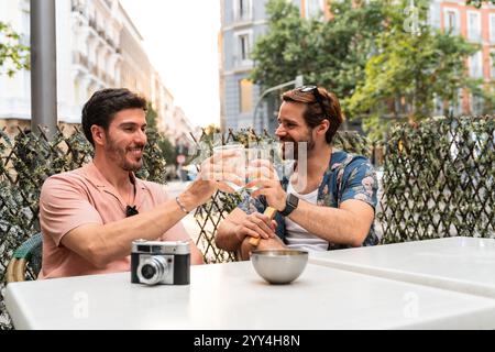 Ein fröhliches schwules Paar toast mit Gläsern Wasser, während es an einem Café-Tisch auf einer Stadtstraße sitzt, flankiert von üppigem Grün, mit einer alten Kamera im Th Stockfoto