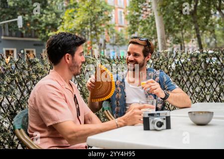 Ein fröhliches schwules Paar teilt sich einen unbeschwerten Moment bei einem Drink in einem Café im Freien, wobei ein Mann einen Ventilator hält und der andere ein Glas Orangengetränk Stockfoto