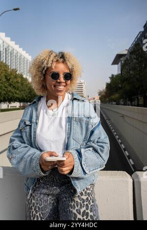Eine junge dominikanische Afro-Frau mit lockigen blonden Haaren, Sonnenbrille und Jeansjacke, lächelt hell, während sie in einem modernen urb ein Smartphone hält Stockfoto