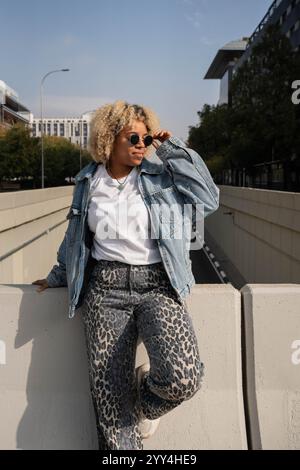 Eine stilvolle junge Frau mit Jeansjacke und Sonnenbrille lehnt sich selbstbewusst an einem städtischen Geländer. Das Stadtbild im Hintergrund ergänzt h Stockfoto