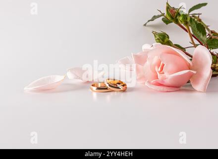 Zwei Eheringe ruhen elegant neben einer zarten rosa Rose und ihren verstreuten Blütenblättern. Der weiche weiße Hintergrund verstärkt die romantische und zeitlose Gebühr Stockfoto