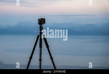 Eine Kamera auf einem Stativ fokussiert auf einen ruhigen See mit majestätischen Bergen in der Ferne. Die ruhige Szene fängt die Ruhe der Natur während du ein Stockfoto