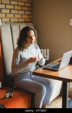 Eine blinde Frau sitzt an einem Holztisch und arbeitet in einem Café mit einem Laptop und Kopfhörern. Sie scheint konzentriert zu sein, mit einem weißen Stock an ihrer Seite, der Technik umschließt Stockfoto