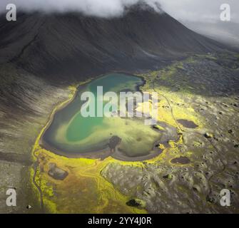 Aus der Vogelperspektive auf einen pulsierenden, herzförmigen vulkanischen Kratersee, der von üppigem Moos, schattigen Bergen und verstreuten Lavafeldern umgeben ist, unter einem stimmungsvollen Clou Stockfoto