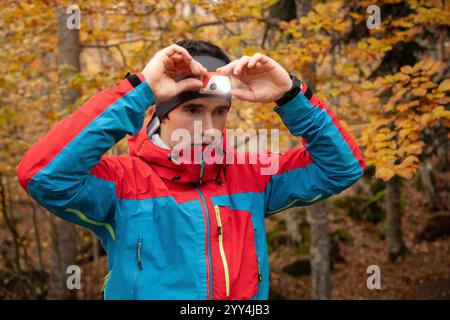 Ein junger Läufer stellt seine Scheinwerfer in einem lebhaften Herbstwald ein. Mit einer farbenfrohen Laufjacke bereitet er sich auf einen Lauf bei Dämmerung oder Dämmerung vor, Highli Stockfoto