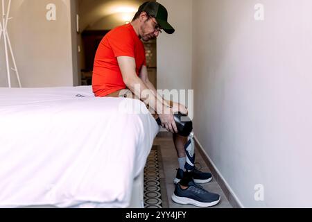 Ein Mann sitzt auf einem Bett und stellt seine Beinprothese in einer einfachen, lässigen Umgebung ein. Er trägt ein rotes Hemd und eine rote Mütze, wobei er sich auf sein Bein konzentriert Stockfoto