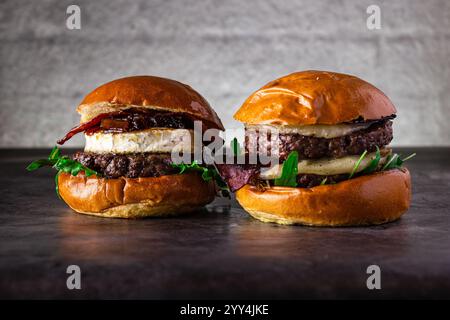 Zwei köstliche Gourmet-Burger mit saftigen Rindfleischpasteten, cremigem Brie-Käse, knusprigem Speck und frischem Rucola, die alle zwischen Hochglanz-Brioche eingelegt werden Stockfoto