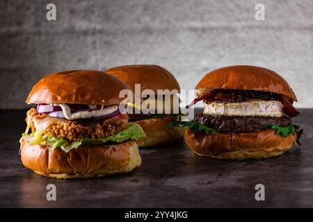 Drei Gourmet-Burger mit saftigen Patties, geschmolzenem Käse, Zwiebeln, knusprigem Salat, Speck, und weiche Brötchen. Präsentiert auf einer dunklen Oberfläche vor einem rustikalen bac Stockfoto