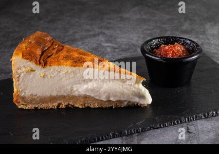 Eine köstliche Scheibe cremiger Käsekuchen liegt auf einer dunklen Schieferoberfläche, begleitet von einem kleinen Gericht mit lebendiger Guavapaste. Perfekt zum Nachtisch oder Essen Stockfoto