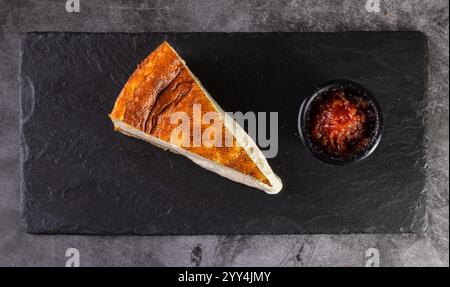 Ein köstliches Stück Käsekuchen mit goldbrauner Oberseite, serviert auf einer Schieferplatte, begleitet von einer Seite mit reichhaltiger Beerensauce in einem kleinen schwarzen Gericht. Pe Stockfoto