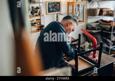 Seitenansicht eines älteren männlichen Besitzers, der während der Arbeit in einem Antiquitätengeschäft den Stuhl repariert Stockfoto