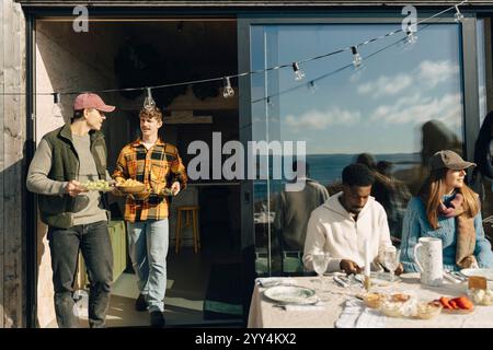 Männliche Freunde halten Essen, während sie an sonnigen Tagen durch die Tür auf der Veranda laufen Stockfoto