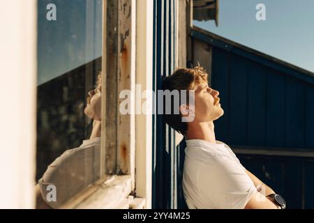 Seitenansicht eines jungen Mannes, der an sonnigem Tag mit geschlossenen Augen an die Tür lehnt Stockfoto