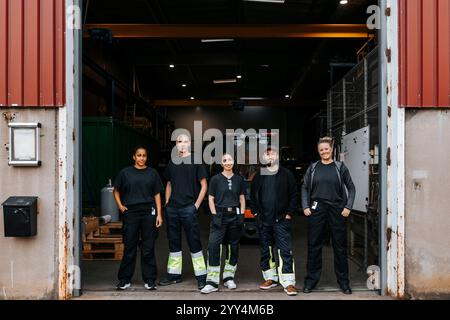 Porträt von lächelnden multirassischen männlichen und weiblichen Lagerarbeitern, die mit Händen in Taschen am Eingang der Fabrik stehen Stockfoto