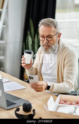 Ein gutaussehender Seniorenmann trinkt Kaffee und überprüft sein Smartphone an einem eleganten Schreibtisch. Stockfoto