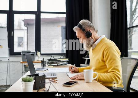 Ein älterer Mann in einem gelben Pullover schreibt an seinem Schreibtisch, schlürft Kaffee und trägt Kopfhörer. Stockfoto