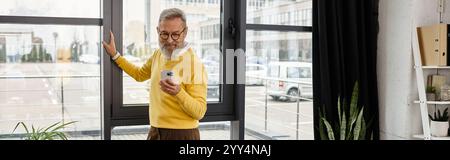 Ein Senior Gentleman in einem gelben Pullover überprüft sein Handy, während er nach draußen blickt, um einen Blick auf die Stadt zu werfen. Stockfoto
