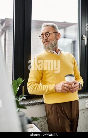 Ein reifer Mann hält eine Kaffeetasse, blickt aus dem Fenster, lässig in einen gelben Pullover gekleidet. Stockfoto