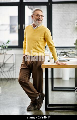 Reifer Mann mit grauem Bart lächelt, während er sich auf einen Schreibtisch in einem hellen Bürobereich lehnt. Stockfoto