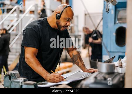 Reifer männlicher Fabrikarbeiter, der im Lager mit einem Laptop über das Headset spricht Stockfoto