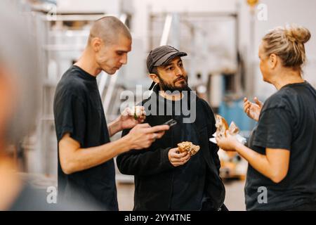 Multirassische männliche und weibliche Lagerarbeiter essen Burger, während sie in der Fabrik miteinander reden Stockfoto