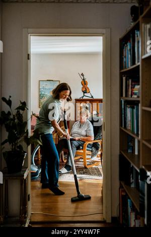 Seniorin sitzt im Wohnzimmer, während eine Pflegeperson zu Hause den Boden saugt Stockfoto