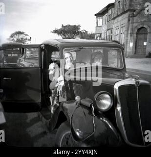 Um die 1940er Jahre, historisch, ein älteres Ehepaar, das in ihrem Morris-Auto der damaligen Zeit sitzt, ein einzelner Scheibenwischer, über der Fahrerseite der Windschutzscheibe auf Dachhöhe befestigt, England, Großbritannien. Teppich auf dem Schoß des männlichen Fahrers. Beachten Sie, dass die Türen in der Mitte aufklappbar sind und sich vom Fahrer in Richtung der Mitte des Fahrzeugs öffnen. Solche Türen wurden allgemein als „Selbstmordtüren“ bezeichnet, da sie gefährlich waren, da man herausfallen könnte, wenn man versehentlich während der Fahrt geöffnet wird. Stockfoto