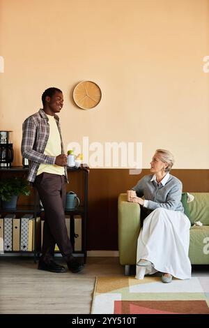 Vertikale Aufnahme einer Reifen Frau in elegantem Outfit, die mit einem jüngeren afroamerikanischen männlichen Kollegen spricht, der auf einer bequemen Couch sitzt, während er im gemütlichen Büro mit beigefarbenen Wänden eine Kaffeepause einlegt Stockfoto