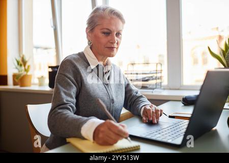 Mittelgroße Aufnahme einer erfahrenen, reiferen Frau, die Notizen auf Papier schreibt, während sie mit dem Laptop am Schreibtisch arbeitet, in einem modernen Büro, im Kopierraum Stockfoto