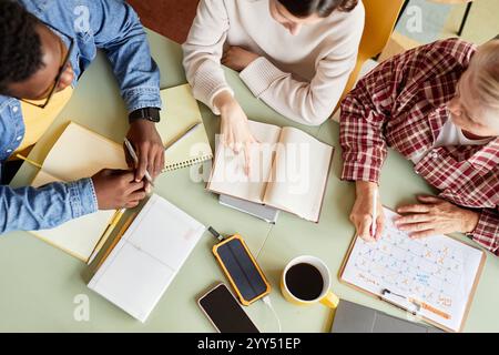 Top-Ansicht eines Teams von Kollegen, die monatlich einen Zeitplan planen, um die Tagesordnung zu besprechen, während sie am Meeting-Tisch sitzen und mit Notizbüchern und Büromaterial am Arbeitsplatz überfüllt sind Stockfoto