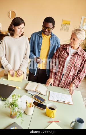 Vertikale Aufnahme des lächelnden modischen Schwarzen Mannes, der mit weiblichen Kollegen die Agenda diskutiert, die am Tisch steht, überfüllt mit Terminplänen und Bürounterlagen in modernen Büros Stockfoto