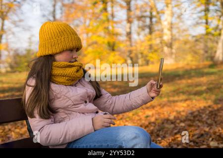 Teenager-Mädchen, das Selfie-Foto-Selbstporträts auf dem Smartphone macht. Kind posiert auf einer Bank im Herbstpark. Lifestyle, soziale Medien. Stockfoto