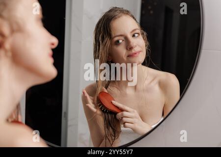 Beauty-Routine. Hübsche Frau Kämmt Ihr Schönes Haar Mit Pinsel, Während Sie Neben Spiegel Im Badezimmer Steht, Attraktive Junge Dame, Die Auf Ihren Reflec Blickt Stockfoto
