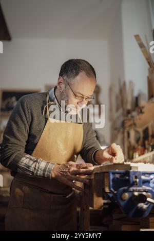 Ein fokussierter Geigenbauer, der in einer gemütlichen Werkstatt an einer Geigenpartie arbeitet und eine Schürze trägt Stockfoto