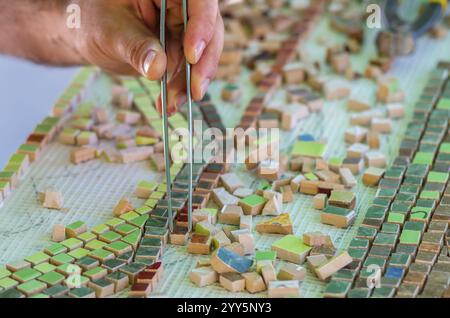 Keramik Design. Keramik Mosaik. Der Mann organisiert mit einer Pinzette ein Puzzle von keramischen Elementen für den Bau von keramischen Kunstwerken. Männer terraco Stockfoto