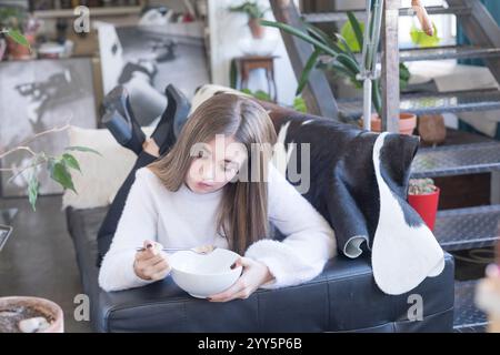 Eine weiße Frau in einem weißen Pullover liegt auf einem Sofa und isst Müsli. Moderne Einrichtung mit Pflanzen und kreativem Dekor sorgt für eine gemütliche, künstlerische Atmosphäre. Stockfoto