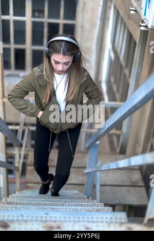 Weiße junge Frau mit langen blonden Haaren, mit Kopfhörern, grüner Jacke und weißem Hemd blickt aufmerksam. Urbane Umgebung im Freien mit sanftem Licht Stockfoto