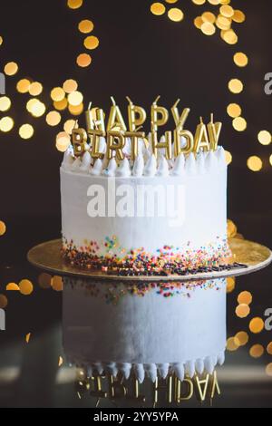 Geburtstagskuchen mit Kerzen, Girlande mit hellen Bokeh-Lichtern auf dem Hintergrund. Der weiße Kuchen ist mit farbigen Streuseln dekoriert und steht auf einem Stockfoto