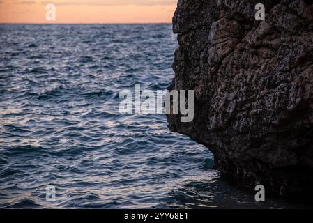Sonnenuntergang an der Küste mit felsigen Klippen. Küste in der Nähe eines Berges mit Felsen und kleinen Wellen. Nahaufnahme des Steinfelsens in klarem blauem Wasser. Stockfoto