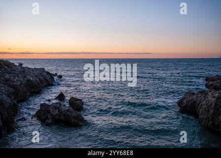 Sonnenuntergang an der Küste mit felsigen Klippen. Küste in der Nähe eines Berges mit Felsen und kleinen Wellen. Nahaufnahme des Steinfelsens in klarem blauem Wasser. Stockfoto