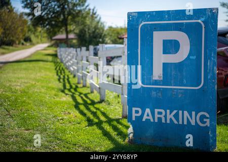 Großes blaues Parkschild, das sich an einen alten hölzernen Weißen Viehzaun lehnt, auf einer Landstraße. Ein Bauernhaus im Hintergrund. Stockfoto