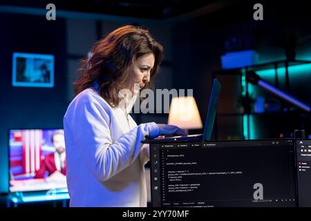 IT-Spezialist, der in einem neonbeleuchteten Zuhause mit Laptop arbeitet, Programme und Systeme testet und implementiert. Software-Techniker überprüft den Code, der auf Computermonitoren angezeigt wird, und steht in der Wohnung Stockfoto